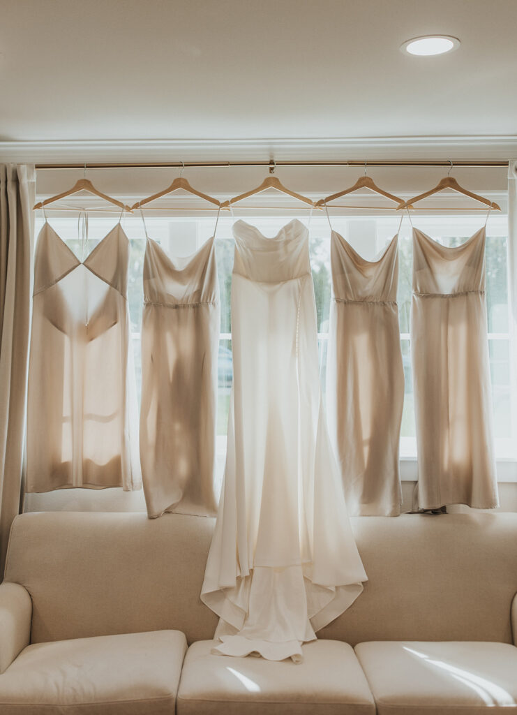 wedding dress and neutral bridesmaid dresses hanging at the window