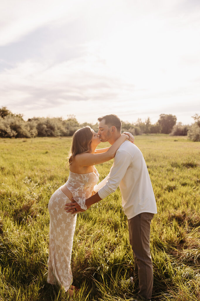 Husband and wife kiss at golden hour in a grassy field while he holds her bump