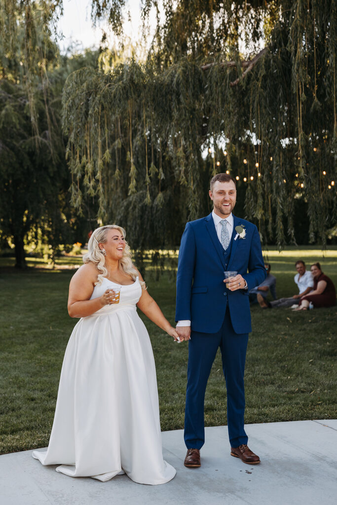 Bride and groom stand holding hands with drink in the other laughing during speeches in the Promise Garden wedding reception space.