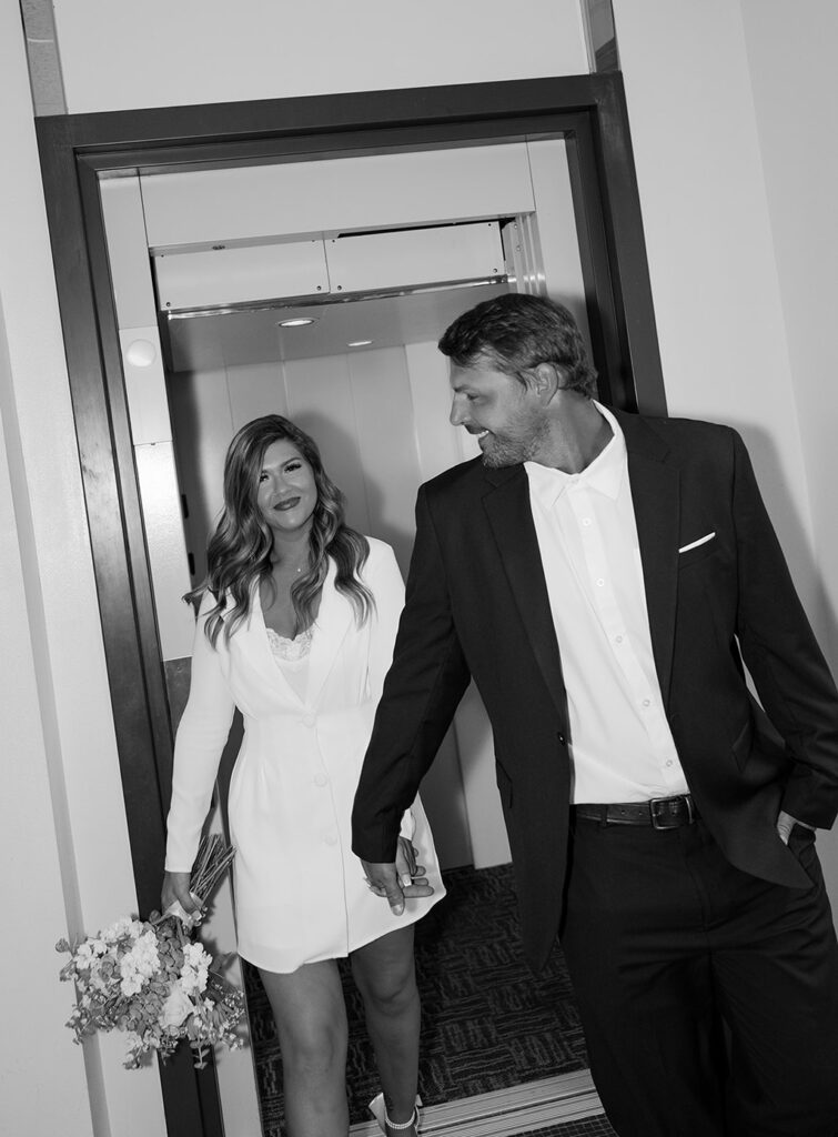 Bride and groom holding hands and walking together indoors, photographed in a black and white editorial wedding photography style.
