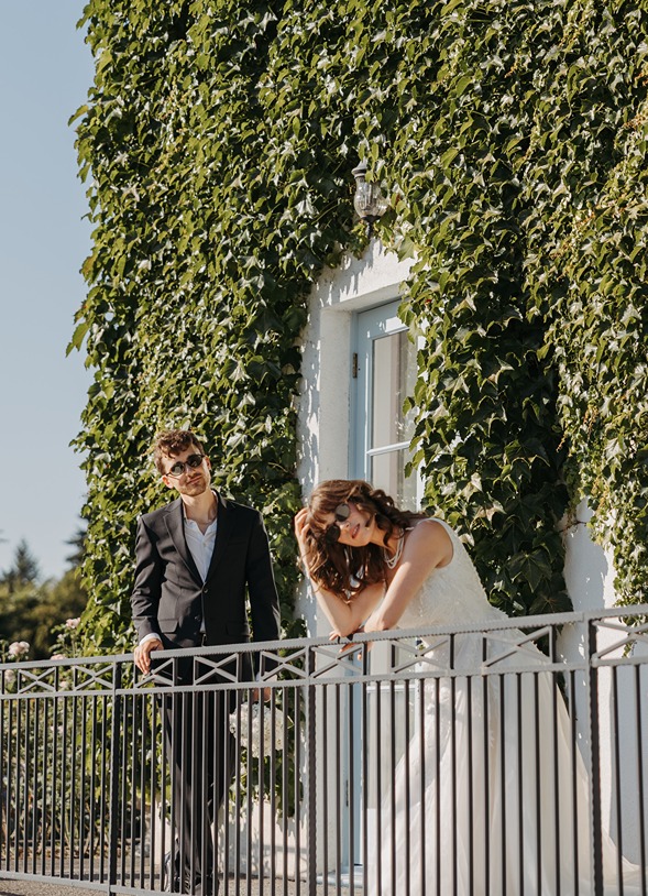 Bride and groom standing on a balcony framed by ivy-covered architecture, photographed by Kat Nielsen Photography in a composed editorial wedding style.