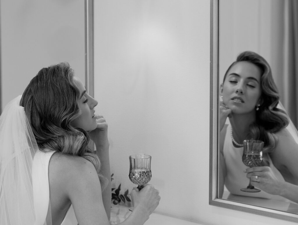 Bride holding a glass and looking at her reflection in a mirror, captured in a black and white editorial wedding photography style.