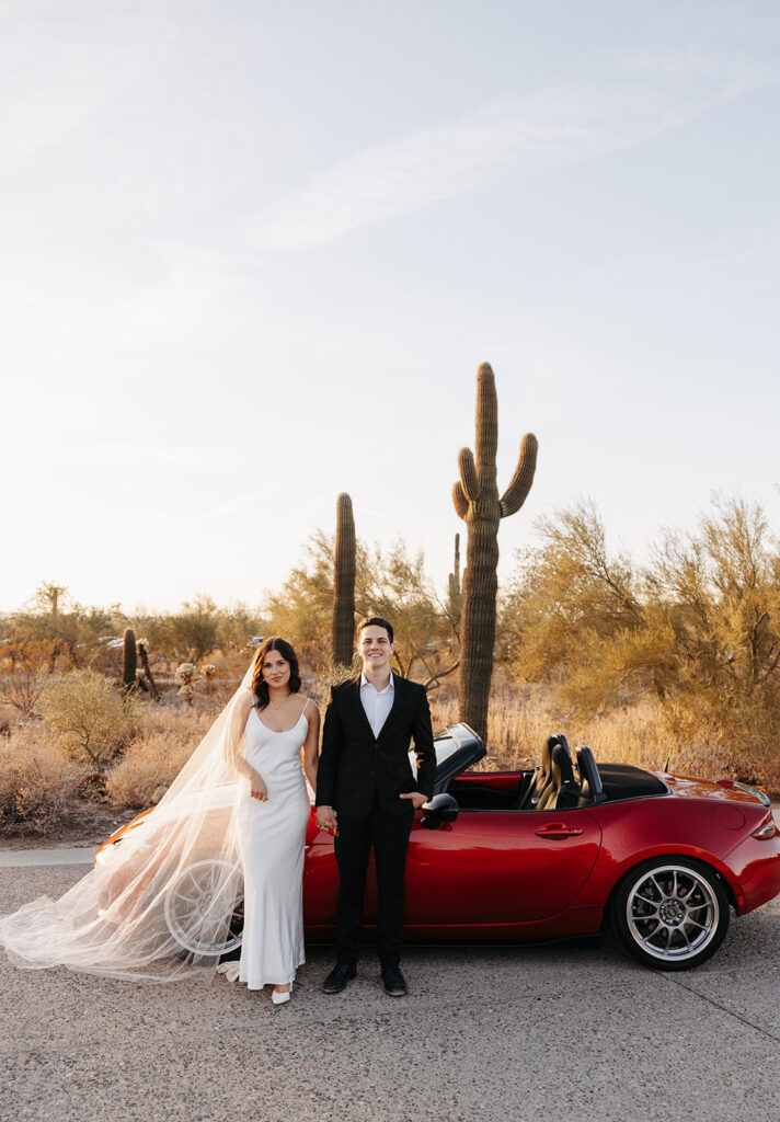 Couple standing hand in hand beside a red convertible in the Arizona desert, surrounded by cacti during golden hour.