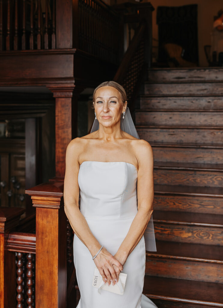 Bride standing on a wooden staircase inside a historic venue, softly lit and composed in a minimalist strapless wedding gown.