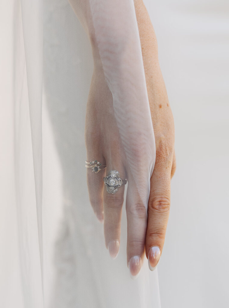 Close-up wedding photograph of a bride’s hand resting softly beneath a sheer veil, showcasing an engagement ring and wedding band in soft natural light.