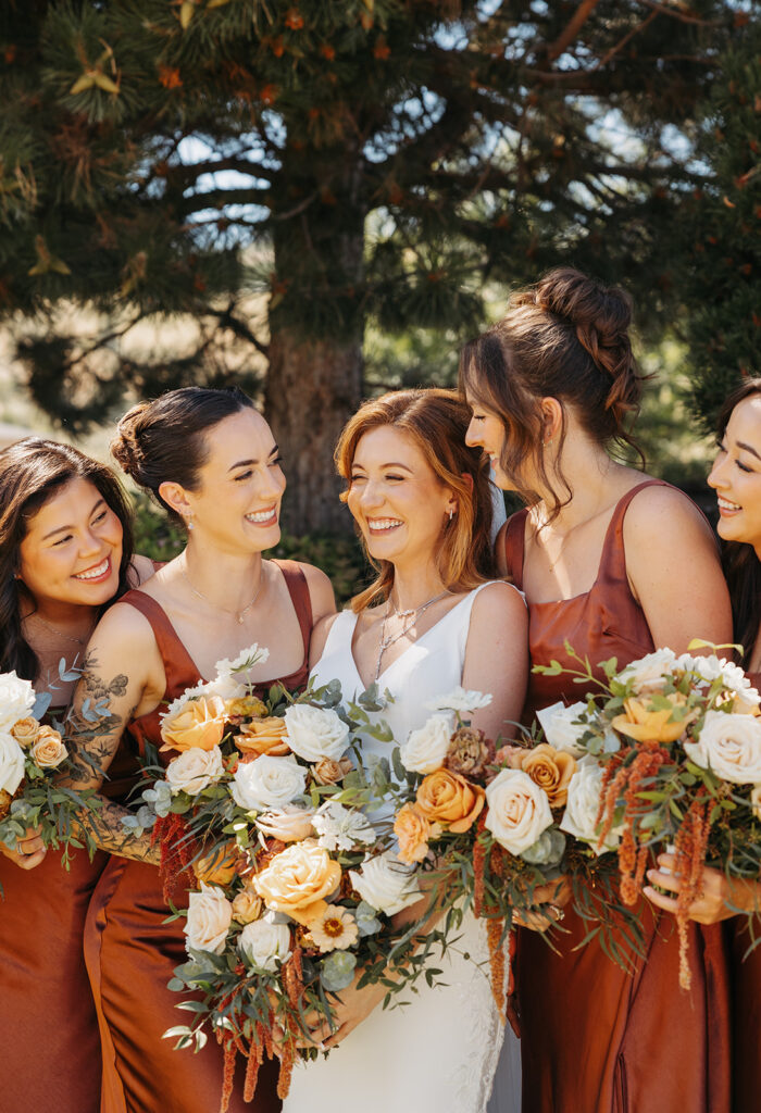 Bride laughing with her bridesmaids while holding lush floral bouquets in warm autumn tones, photographed outdoors in soft natural light.
