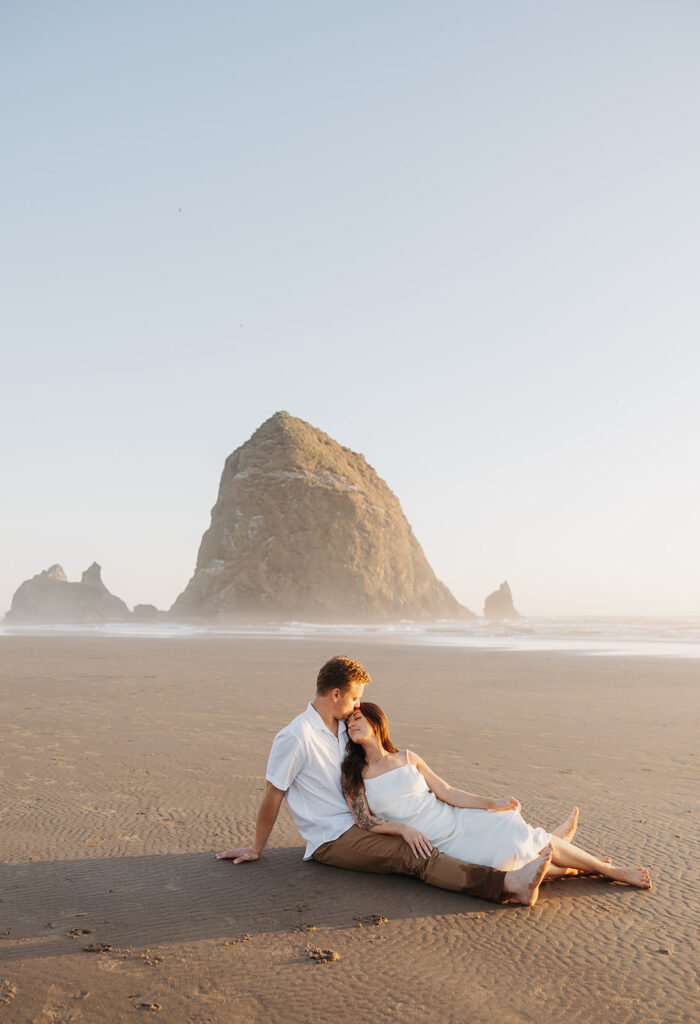 Couple sitting together on a beach with Haystack Rock in the background, photographed in an editorial wedding photography style.