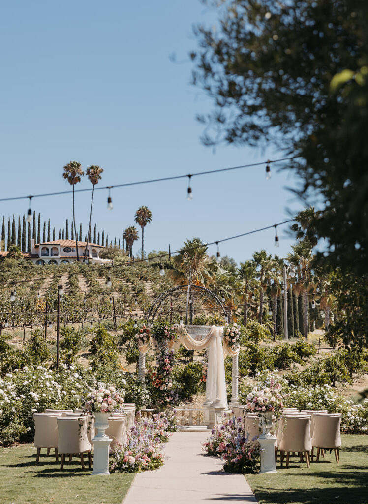 European-inspired outdoor wedding ceremony set among vineyards in Temecula, California, featuring floral-lined aisle, draped ceremony arch, and soft natural light.