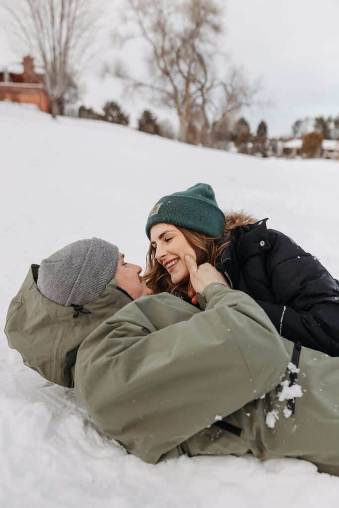 Couple laughing together in the snow, bundled in winter jackets and beanies, sharing a candid, intimate moment outdoors.