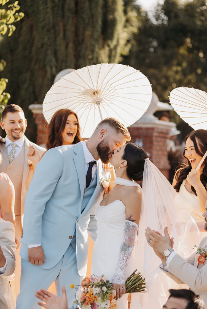 Bride and groom sharing a kiss at Oakshire Estate, surrounded by guests holding white parasols, highlighting lace sleeves, veil layers, and soft evening light.