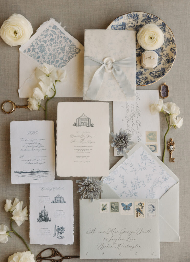 Minimalist wedding stationery flat lay designed by Events by Edz featuring soft blue and ivory paper goods, vintage stamps, delicate florals, and refined textures. Photographed by Kat Nielsen Photography