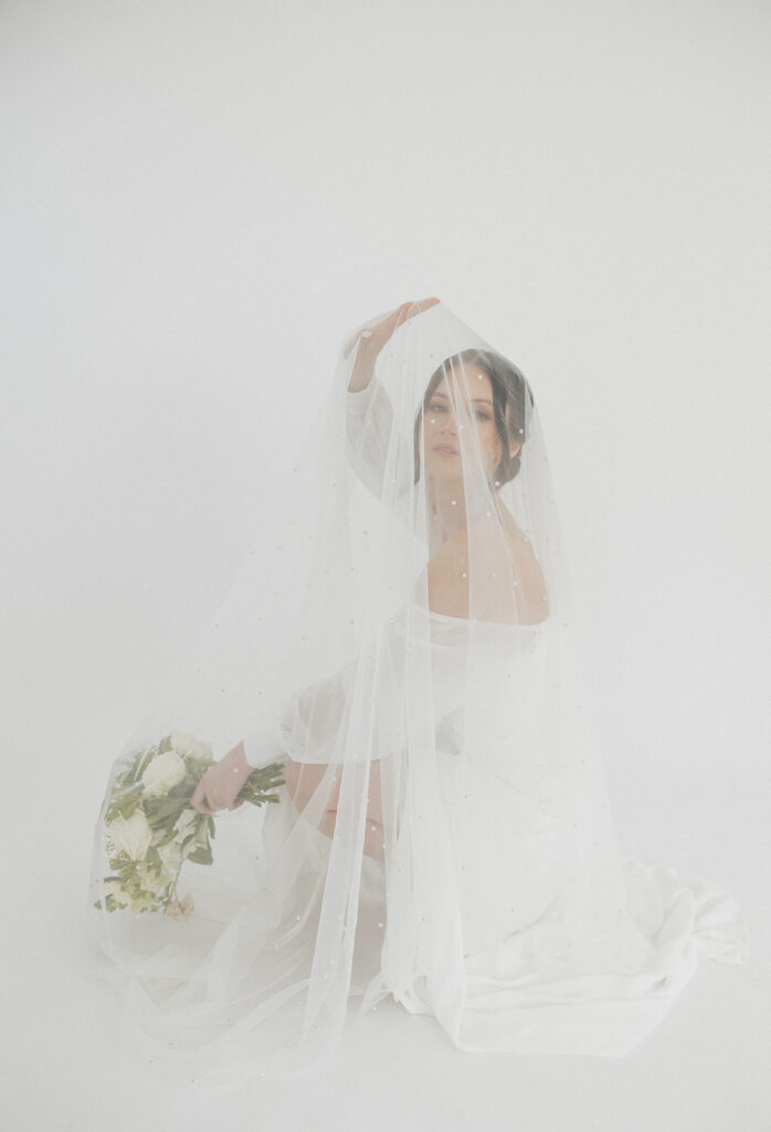 Bride draped in a sheer veil holding a bouquet, surrounded by soft white space and diffused light, creating an ethereal and minimalist portrait.