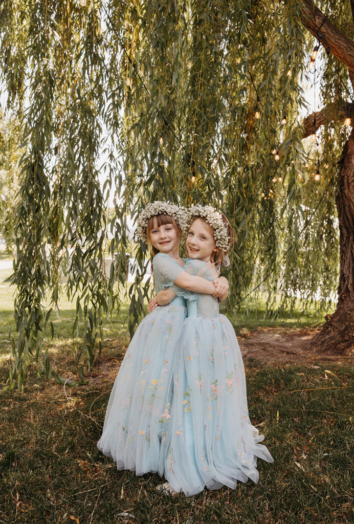 Two flower girls in embroidered blue tulle dresses with floral crowns standing beneath a willow tree, highlighting layered fabric textures and soft natural light.