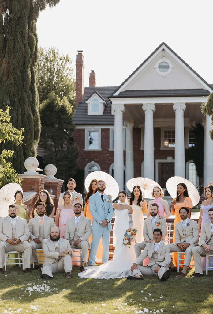 Bride and groom surrounded by their wedding party in front of the Oakshire Estate mansion, with white parasols, pastel attire, and soft evening light.